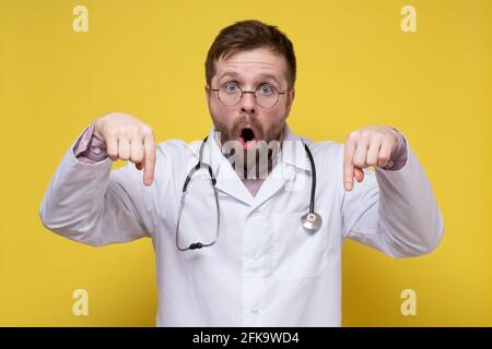 Aufgeregter Arzt in einem weißen Mantel mit einem Stethoskop um den Hals zeigt mit Zeigefinger nach unten, sieht empört mit offenem Mund. Stockfoto