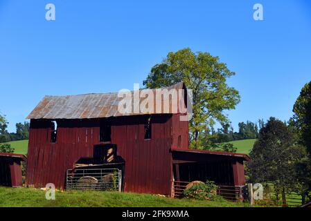 Rusti, eine rote Holzscheune, liegt auf einem Hügel im Osten von Tennessee. Es ist zweistöckig mit einem Loft. Im Eingang zur Scheune sitzen runde Heuballen. Tennessee Hill Stockfoto