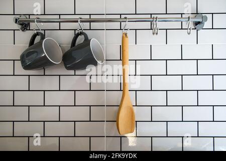 Nahaufnahme des Küchenzimmers mit Holzlöffel und grauer Kaffeetasse, die an einer Stahlschiene an einer weißen Ziegelwand hängen, Innenausstattung und Wohneinrichtung in modernem Stil Stockfoto