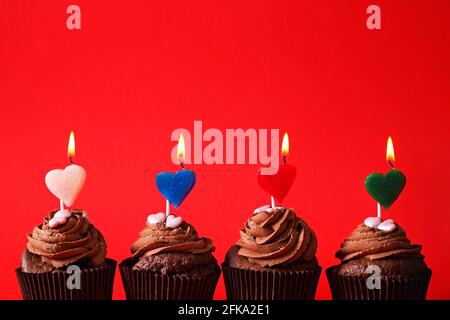 Vier köstliche Schokolade Butterbecher Cupcake, brennendes Herz Form festliche Kerzen, Zucker Belag, isoliert auf rot. Kakao Bäckerei, rustikale Wand Hintergrund, Stockfoto