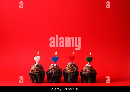 Vier köstliche Schokolade Butterbecher Cupcake, brennendes Herz Form festliche Kerzen, Zucker Belag, isoliert auf rot. Kakao Bäckerei, rustikale Wand Hintergrund, Stockfoto