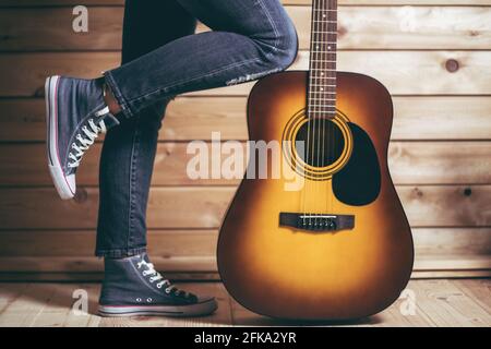 Akustische sechssaitige gelb-braune Gitarre und weibliche Beine in Jeans, in der Nähe einer Holzwand Stockfoto