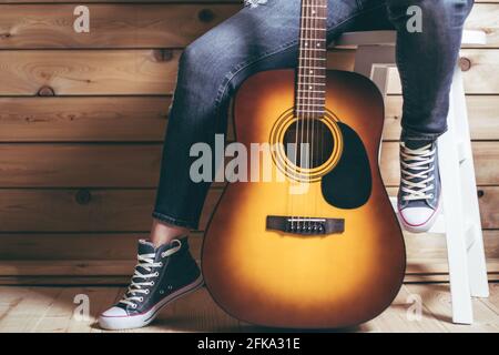 Akustische sechssaitige gelb-braune Gitarre und Beine einer Frau, die auf einem Hocker in Jeans sitzt, in der Nähe einer Holzwand Stockfoto