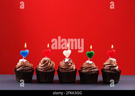 Köstliche Schokolade Butterbecher Cupcakes, brennendes Herz Form festliche Kerzen, Zucker Belag, Beton Textur Tabelle. Kakaobäckerei, rustikaler grauer Kaffee s Stockfoto