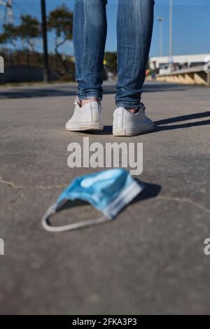 Die Maske liegt auf dem Boden mit den Füßen einer Frau in weißen Turnschuhen und Jeans, die auf die neue Normalität nach dem Impfstoff zugeht, Ende der Pandemie, neue Normalität, Ende der Covid-19. Stockfoto