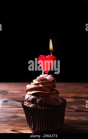 Köstliche dunkle Schokolade Butterbecher Cupcakes, brennende Herzform Kerze Belag, Holz strukturierte Tisch. Kakao-Bäckerei, rustikale rote Keramikplatte Kaffee Stockfoto