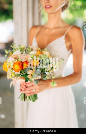 Die Braut steht zwischen weißen, mit grünen Reben umflochtenen Säulen und hält ein Hochzeitsstrauß mit orangen Butterblumen aus nächster Nähe Stockfoto