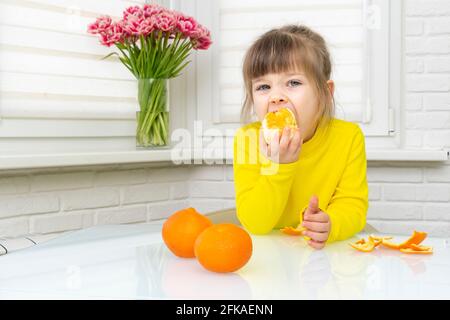 Glückliches kleines Mädchen in gelben Kleidern, das eine große Mandarine isst Ganze, während man an einem Tisch in einer weißen Küche sitzt Stockfoto