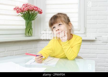 Kleines kaukasisches Mädchen in gelben Kleidern sitzt an einem Tisch In einem weißen Raum und zieht Stockfoto