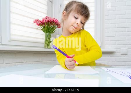 Das kleine Mädchen sitzt an einem Tisch in einem weißen Raum und zeichnet. Stockfoto