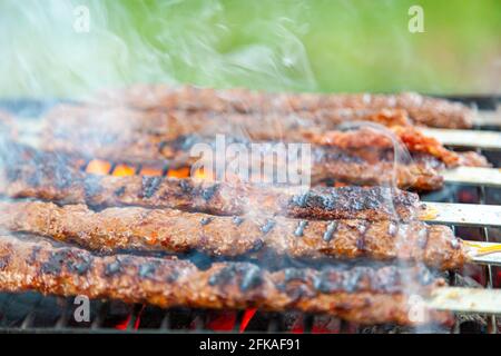 Beim Kochen köstlicher türkischer Adana Kebab auf einem tragbaren Holzkohlegrill an einem Picknickplatz kommt Rauch und Flamme aus dem brutzelnden Fleisch. Koftes sind li Stockfoto