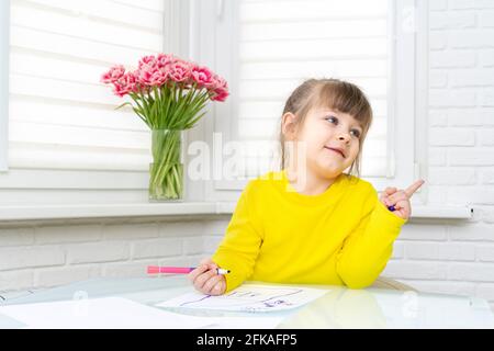 Das kleine Mädchen sitzt an einem Tisch in einem weißen Raum und zeichnet. Stockfoto