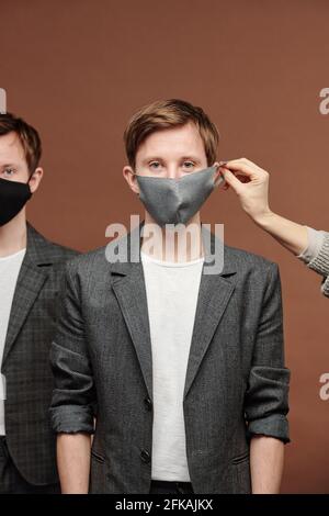 Hand Putting Maske auf dem Ohr zu jungen Kerl in grau Anzug steht mit Zwillingsbruder auf braunem Hintergrund Stockfoto