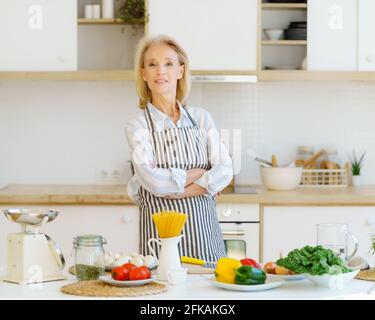 Porträt einer schönen, positiv reifen Frau in der Schürze in der Küche, die die Kamera ansah und lächelte, einer pensionierten Großmutter, die neben dem Tisch mit gesunden Zutaten stand, während sie für die Familie zu Hause kochte Stockfoto