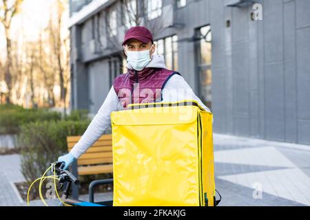 Junger Kurier in medizinischer Maske liefert Lebensmittel mit gelbem Thermalrucksack, fährt mit dem Fahrrad in der Stadt. Konzept für den Lieferservice von Lebensmitteln Stockfoto