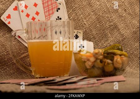 Spielkarten, ein Becher mit leichtem, ungefiltertem Bier und eine Glasschale mit Gurken und Pilzen auf einem Tisch, der mit grobem Tuch bedeckt ist. Nahaufnahme, selektiver FOC Stockfoto