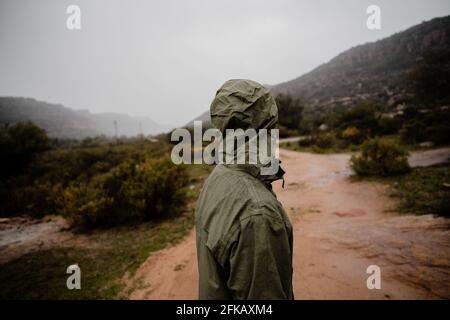 Junger, fittlicher Läufer, der sich vom Laufen auf einem Kiesberg ausruhte Weg im Regen mit Regenjacke bewölktes Wetter grüne Büsche Stockfoto