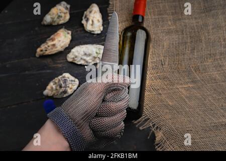 Hand mit Austernmesser in Schutzhandschuhen. Nahaufnahme. Austern und Weinflasche im Hintergrund Stockfoto