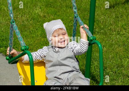 Das kleine kaukasische Mädchen, 2 Jahre alt, schwingt auf einer Schaukel und lacht fröhlich. Fröhliches Kind in einem grauen Hut spielt auf dem Spielplatz, Nahaufnahme Porträt Stockfoto