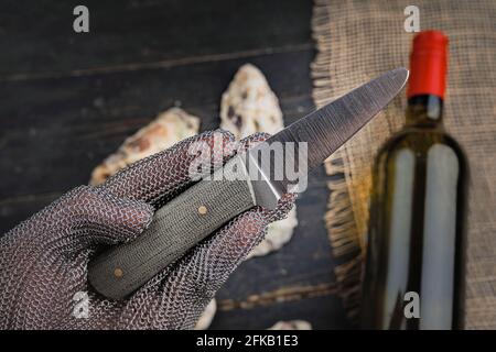 Hand mit Austernmesser in Schutzhandschuhen. Nahaufnahme. Austern und Weinflasche im Hintergrund Stockfoto