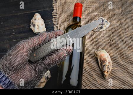 Hand mit Austernmesser in Schutzhandschuhen. Nahaufnahme. Austern und Weinflasche im Hintergrund Stockfoto