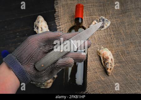 Hand mit Austernmesser in Schutzhandschuhen. Nahaufnahme. Austern und Weinflasche im Hintergrund Stockfoto