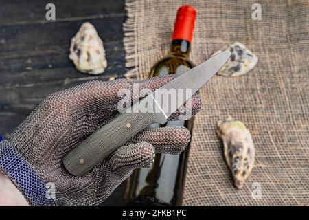 Hand mit Austernmesser in Schutzhandschuhen. Nahaufnahme. Austern und Weinflasche im Hintergrund Stockfoto