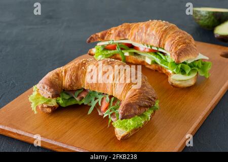 Appetitliche französische Croissant-Sandwiches mit Lachs, Avocado, Spinat und Salat, serviert auf einem Holzschreibtisch auf grauem Hintergrund. Leckeres Essen für bre Stockfoto