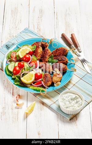 Lamm-Kofta-Kebabs aus gemahlenem Lamm, serviert auf einem blauen Teller mit Joghurtdressing, frischem Gemüsesalat und Zitronenkeilchen, griechische Küche auf einem weißen Holz Stockfoto