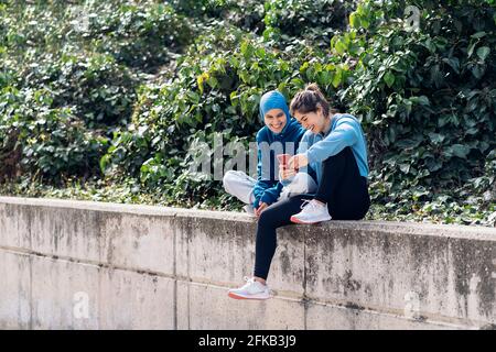 Fröhliche muslimische Frau in Hijab- und Sportkleidung, die mit ihrem Freund auf der Straße sitzt und ein Mobiltelefon benutzt. Stockfoto