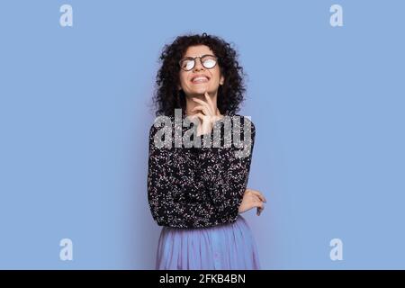 Monochromes Foto einer kaukasischen lockigen Frau mit Brille Und modisches Kleid, das an einer blauen Wand posiert Stockfoto