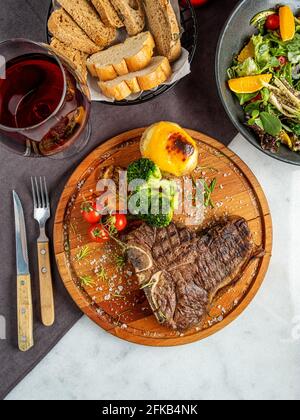 Mittelseltenes gegrilltes T-Bone Steak mit Kartoffelkeilen, Brokkoli und Wein auf Servierbrett Block auf dunklem Hintergrund Stockfoto