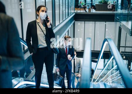 Geschäftsfrau, die über ein Smartphone spricht, während sie sich auf der Rolltreppe nach oben bewegt Während einer Pandemie Stockfoto