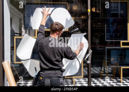 Am 27. April 2021 klebt ein männlicher Retailler-Unternehmer einen Aufkleber auf der Fensterfläche der Jermyn Street-Filiale von Charles Tyrwhitt in London, England. Jetzt, da ihre Filialen nach Pandemiesperrungen wieder geöffnet werden, bietet der Herrenmode-Einzelhändler Charles Tyrwhitt einen Recycling-Service für alte Hemden an. Stockfoto