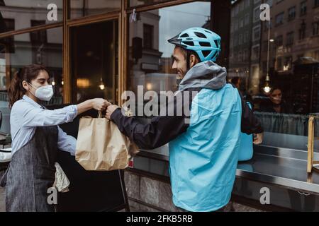 Weibliche Besitzerin, die Paketbestellung an den Lieferer vor dem Deli gab Während einer Pandemie einkaufen Stockfoto