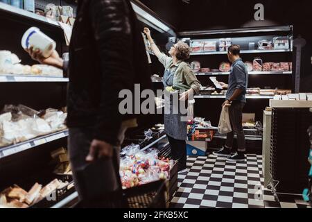 Männliche Kunden sammeln Paketprodukte, während weibliche Besitzer dabeistehen Essen im Regal im Delikatessenladen arrangieren Stockfoto