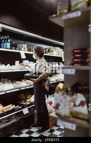 Weibliche Besitzerin, die Lebensmittel auf dem Regal im Deli-Shop arrangiert Während einer Pandemie Stockfoto