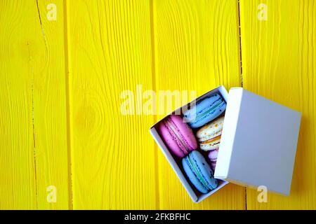 Französische Makronen-Süßigkeiten, verschiedene Geschmacksrichtungen und Farben. Box mit Makronen auf gelbem Holz Holzstruktur Tisch Hintergrund. Geburtstag valentinstag Frühlingsgeschenk Stockfoto