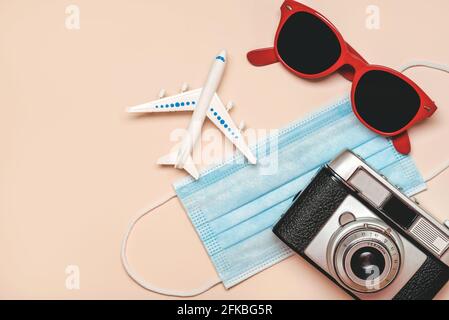 Schützende OP-Maske mit Sonnenbrille, Retro-Fotokamera und Flugzeug mit Platz auf beigem Hintergrund kopieren.Reisekonzept mit covid-19 Stockfoto