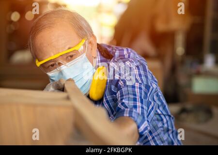 Ältere Arbeiter Holzholzarbeiten Ruhestand Hobby für einen guten Ruhestand, asiatische männliche reifen professionellen Meister der Herstellung von Holzmöbeln mit Gesichtsmaske Protecti Stockfoto