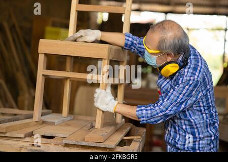 Ältere Arbeiter Holzholzarbeiten Ruhestand Hobby für einen guten Ruhestand, asiatische männliche reifen professionellen Meister der Herstellung von Holzmöbeln mit Gesichtsmaske Protecti Stockfoto