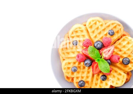 Herzförmige belgische Waffeln mit verschiedenen Beerenmix, Erdbeere, Heidelbeere, Himbeere und rote Johannisbeere, verziert mit Minzblättern auf weißer Keramikplatte Stockfoto