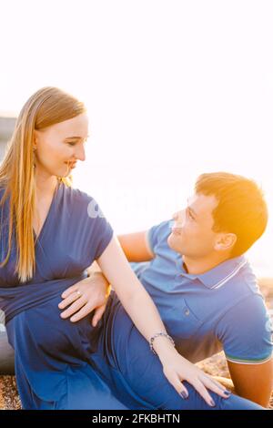 Der Mann hält seine Hand auf den Bauch eines Lächelns Schwanger Frau in einem langen blauen Kleid, während sie auf liegt Der Sand Stockfoto