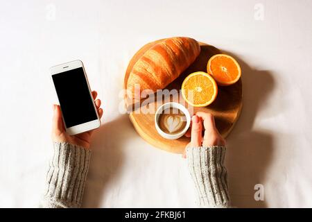 Über dem Kopf geschnittene Aufnahme einer Frau, die ein Mittagessen mit Croissants und Kaffee hatte und ihr Telefon scrollte. POV. Stockfoto