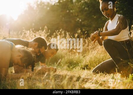 Fitnesstrainer überprüft die Zeit, während männliche und weibliche Athleten trainieren Im Park an sonnigen Tag Stockfoto