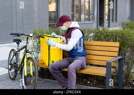 Junger Kurier in medizinischer Maske liefert Lebensmittel mit gelbem Thermalrucksack, fährt mit dem Fahrrad in der Stadt. Konzept für den Lieferservice von Lebensmitteln Stockfoto