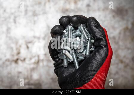 Person, die einen Schutzhandschuh trägt und eine Handvoll Schrauben gegen sie hält Metallhintergrund Stockfoto