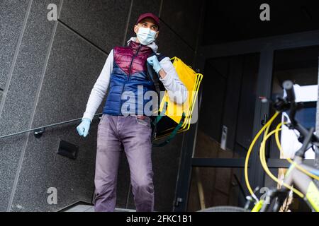 Junger Kurier in medizinischer Maske liefert Lebensmittel mit gelbem Thermalrucksack, fährt mit dem Fahrrad in der Stadt. Konzept für den Lieferservice von Lebensmitteln Stockfoto