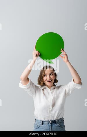 Fröhliche junge Frau in weißem Hemd mit grüner Sprechblase Über Kopf isoliert auf grau Stockfoto
