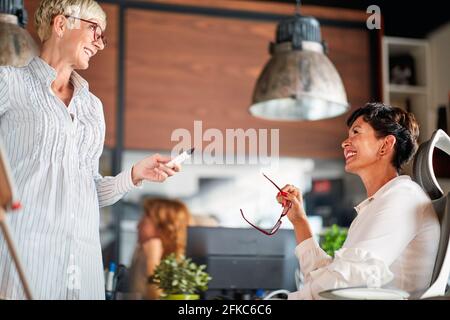 Eine ältere, fröhliche Geschäftsfrau, die in einer angenehmen Atmosphäre am Arbeitsplatz über eine Präsentation mit ihrer Kollegin spricht. Unternehmen, Büro, Job Stockfoto